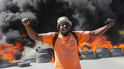 Man yells in front of a wall of burning tyres