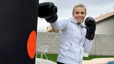 Lara Lewington wears black boxing gloves and punches a punchbag with a orange spot on it.