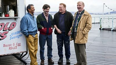 Phaldut Sharma, Iwan Rheon, Mark Lewis Jones and Steffan Rhodri stand on a pier beside a van selling hot food including bacon and egg rolls and jumbo sausages