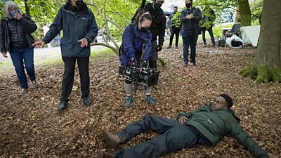 Behind the scenes image of DC Davis Lindo (ROMARIO SIMPSON) on the ground of a wood curreounded filming crew