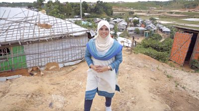 Esmat wears a white and blue dress and headscarf. She stands in front of the tarpaulin-and-metal shelters of a Rohingya refugee camp in Cox's Bazar.