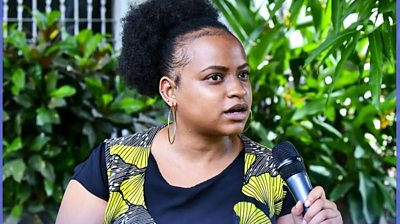 A young woman sits and speaks into a microphone. She is wearing a black T shirt and yellow vest and is sitting in front of a row of plants.