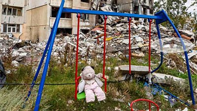 A teddy bear abandoned on a swing next to a destroyed building in Ukraine