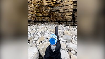 beach covered in boulders with boy in a blue winter hat and water proof jacket pointing to a layered rock cliff face