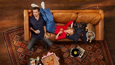 aerial shot of a man sitting on a sofa with a woman laying down and her legs resting on the back of the sofa. A dog on the sofa with a cone on its head