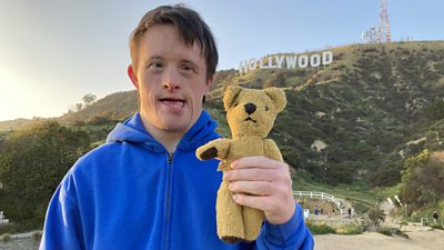 Tommy Jessop stands at the bottom of a hill with the Hollywood sign in the background. He holds a teddy bear.