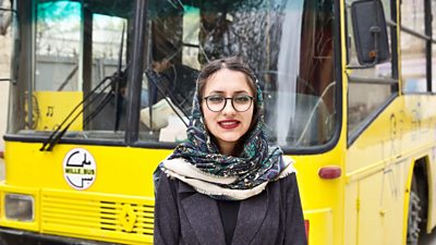 Freshta is wearing a grey coat and patterned headscarf and stands in front of a yellow bus in Afghanistan