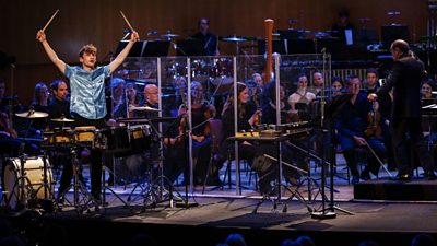 Jordan Ashman holding drum sticks up and standing in front of drum with orchestra behind him