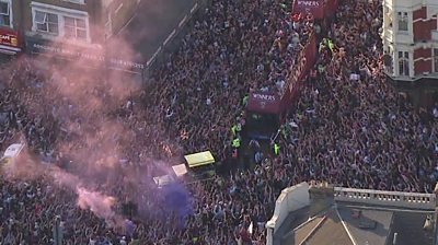 Two buses drive through huge crowds in the street with flares lit