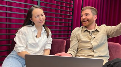 A confused looking woman and a smiling man, sat on a sofa with a laptop