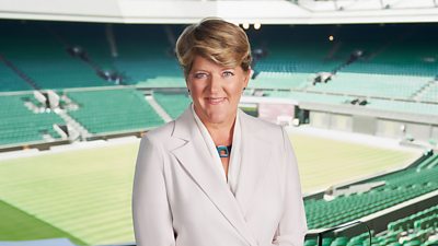 Clare Balding stands in the stands at Wimbledon, wearing a white suit and smiling for the camera