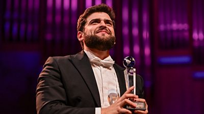 Adolfo Corrado wears a black tuxedo and white shirt and bowtie, he is holding a trophy