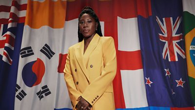 Anita Asante wears a yellow blazer and stands in front of a wall of flags