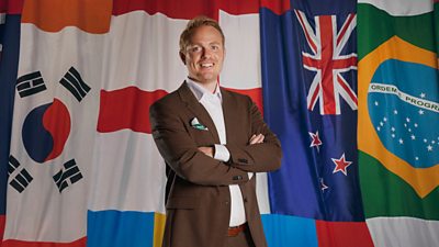 Jonas Eidevall wears a brown suit and is standing with his arms folded in front of a wall of flags