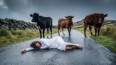 Ruth Wilson as Lorna. She lies on a wet road with three cows standing over her.