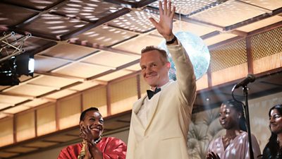 Rob waves across a dance floor wearing a white tuxedo