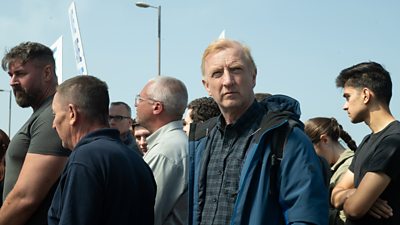 Steffan Rhodri as Geoff - he stands in a crowd of men outdoors, wearing a shirt and coat