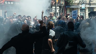 An angry crowd clash with ;police on a street - one person holds a placard which says We Are One