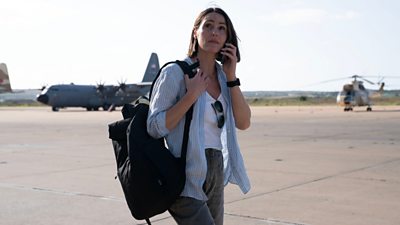 Amy Silva (Suranne Jones) stands on the airbase with her phone by her air, and a place and a helicopter in the background