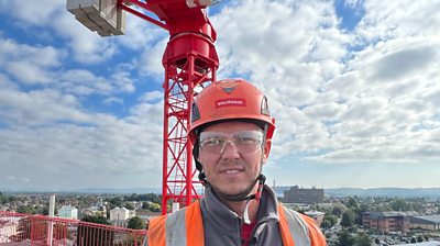 Man smiles with builder hard hat on next to massive red crane
