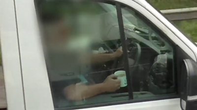 Motorist sat in driver's seat clutching cup of tea with his right hand and steering wheel with his left