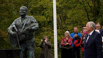 Max Boyce in a crowd standing by a Max Boyce statue