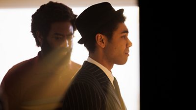 Character Dante Williams wearing pinstripe suit and hat facing the right. Character Deuce Williams in background with sun glare looking slightly down 