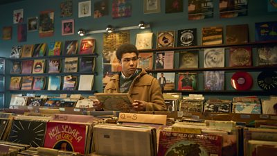 Levi Brown as Dante Williams in a record shop looking to the right at something offscreen