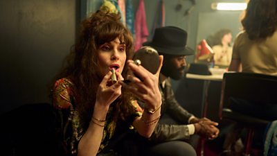 Estella played by Michelle Dockery  putting lipstick on looking in a cosmetic mirror. Deuce Williams played by Nicholas Pinnock in background with hands together looking ahead of him