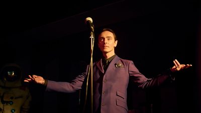 Robbie Carmen played by David Dawson standing slightly back from a microphone in a plum suit with his arms outstretched