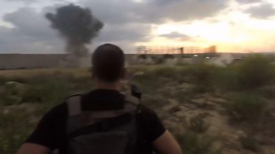 Man running through field as explosions seen in background