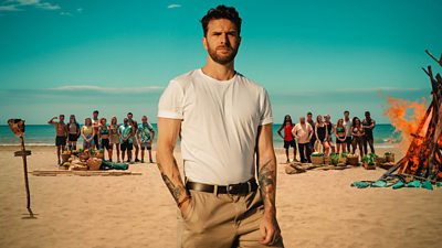 Joel Dommett and the cast of Survivor are pictured on a beach. The contestants stand in two tribes behind him on the sand with provisions in front of them. To the right, a wood fire burns.