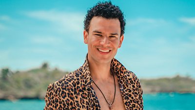 Survivor contestant Christopher stands on the beach wearing an open leopard print shirt, smiling