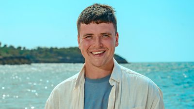 Survivor contestant Matthew stands on the beach wearing a cream shirt over a blue t-shirt, smiling with the sea in the background