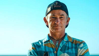 Survivor contestant Pegleg wears a patterned short sleeved shirt and a backwards baseball cap, standing with arms folded on the beach