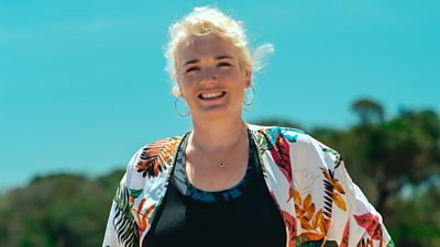 Survivor contestant Rachel wears a white shirt with a multicoloured leaf pattern, over a black top and stands on the beach in the sun