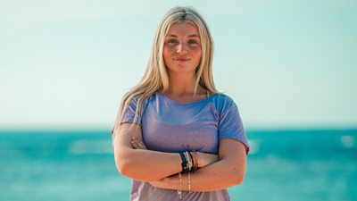 Survivor contestant Rach wears a purple t-shirt and stands with arms folded in front of the sea on the beach