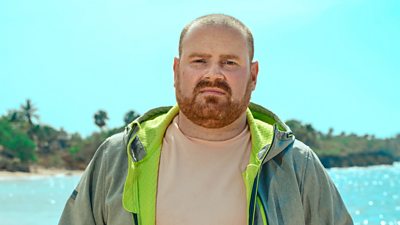 Survivor contestant Richard wears a green jacket over a t-shirt and stands on the shoreline