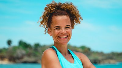 Survivor contestant Sabrina wears a light blue tank top and stands with arms crossed in front of the sea