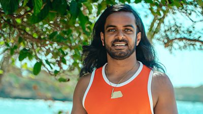 Survivor contestant Shai wears an orange tank top and stands under a tree on the shoreline