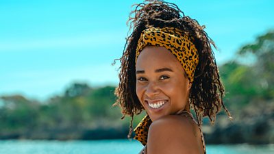 Survivor contestant Tinuke wears a leopard print top and shorts and a leopard print hair wrap, smiling for the camera on the beach