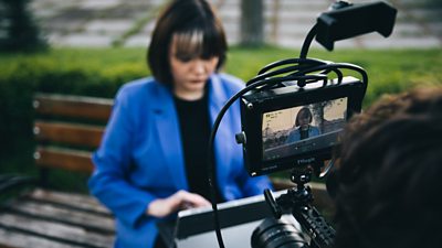 A woman with dark hair wearing a bright blue jacket looks down at her laptop. A camera operator is setting up a camera in front of her.