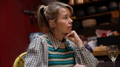 Anna Maxwell Martin sits at a table in character, resting her chin on one hand. There's a small glass of wine in front of her. She looks concerned.