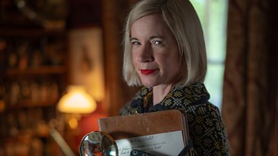 Lucy Worsley holding a brown leather folder with a piece of paper that says Study in Scarlett and she's holding a magnifying glass