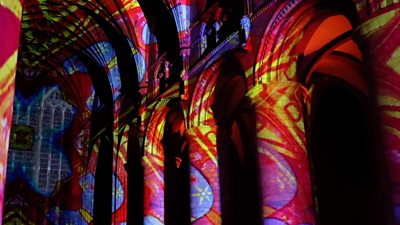 A light display of reds, blues, yellows and purples is projected onto the pillars of Gloucester Cathedral