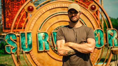 Joel Dommett stands in the sun in front of a large metal disc, featuring the word Survivor in large green block capital letters