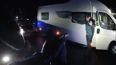Armed police point a gun at a man as he emerges from a motorhome with his hands up.