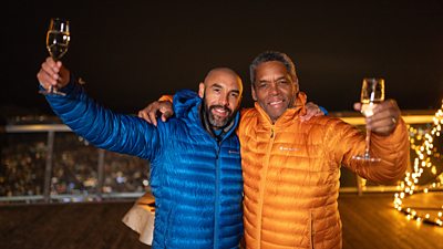 Alex and Noel Beresford raise glasses to celebrate their Celebrity Race Across The World win. They wear winter jackets - Alex's is blue and Noel's is yellow - and pose with an arm around each other in the dark, with twinkling fairy lights and city lights in the background.