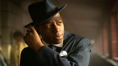 Man looking at something off screen touching the rim of hit hat and dressed in a suit