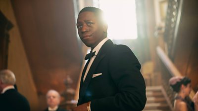 Character Luke Fitzwilliam wearing tuxedo in front of large staircase looking pensive, appears to be a party in the background